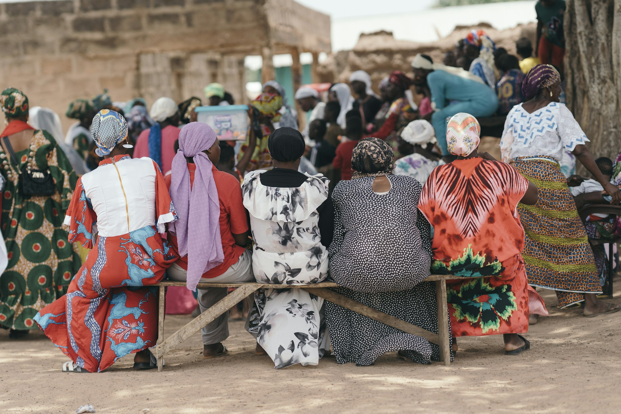 A row of women in colorful traditional dress sit together on a bench at a community gathering in a village in Ghana, with a larger crowd gathered behind them