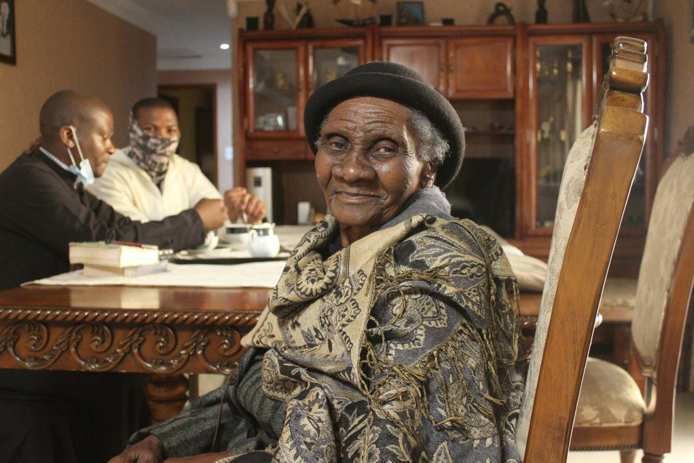 An elderly woman in a hat and patterned shawl smiles warmly at her dining table in a home in Soweto, South Africa, with two visitors seated in the background