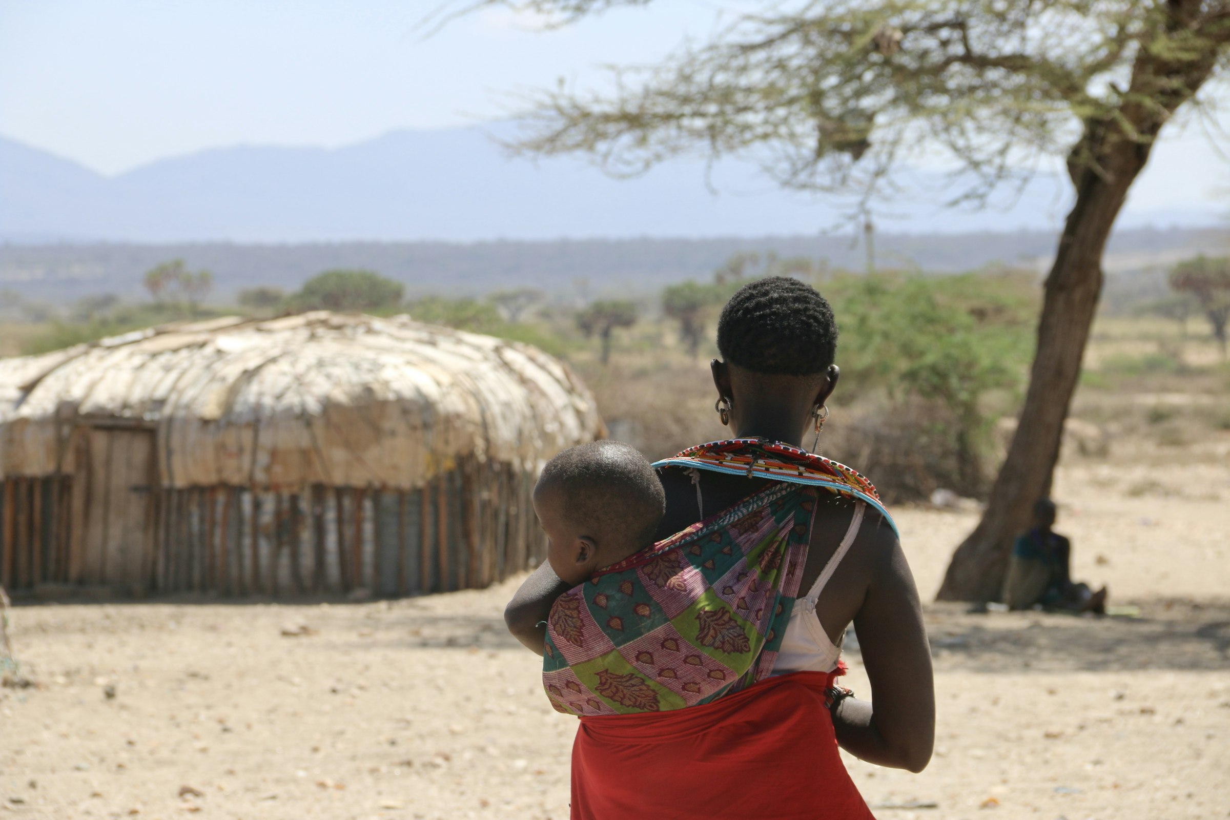 Samburu mother in beaded dress carrying her child past a traditional homestead, Kenya
