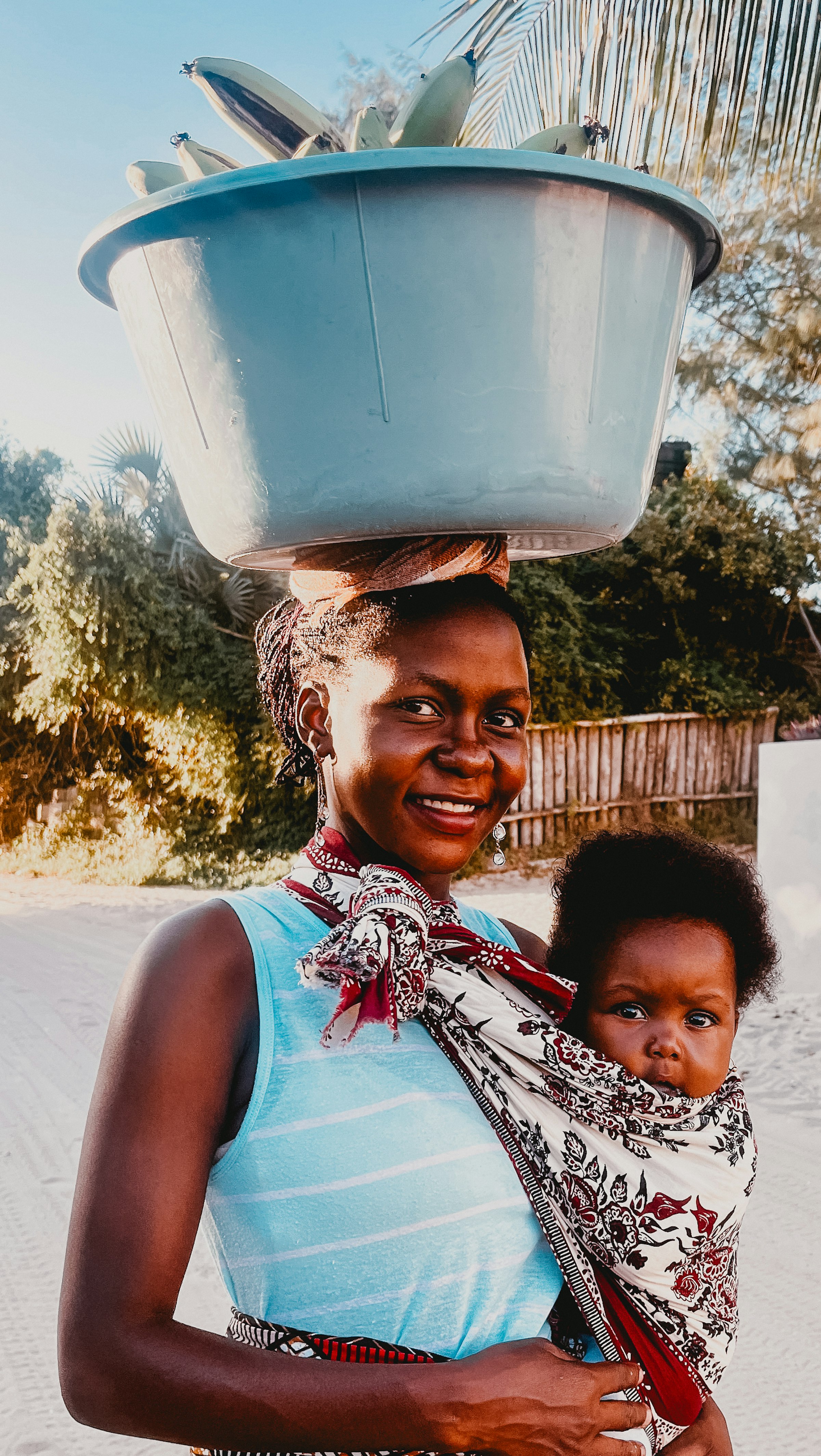 Smiling mother in Mozambique carrying her baby in a cloth wrap, balancing a basin on her head