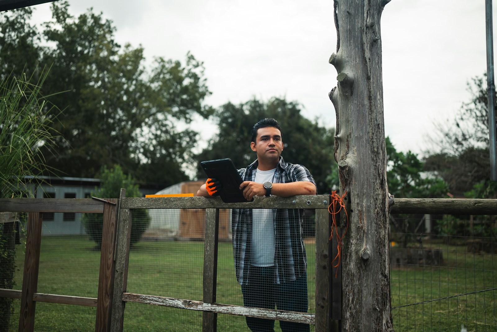 A field worker in gloves holding a tablet outdoors near a wooden fence