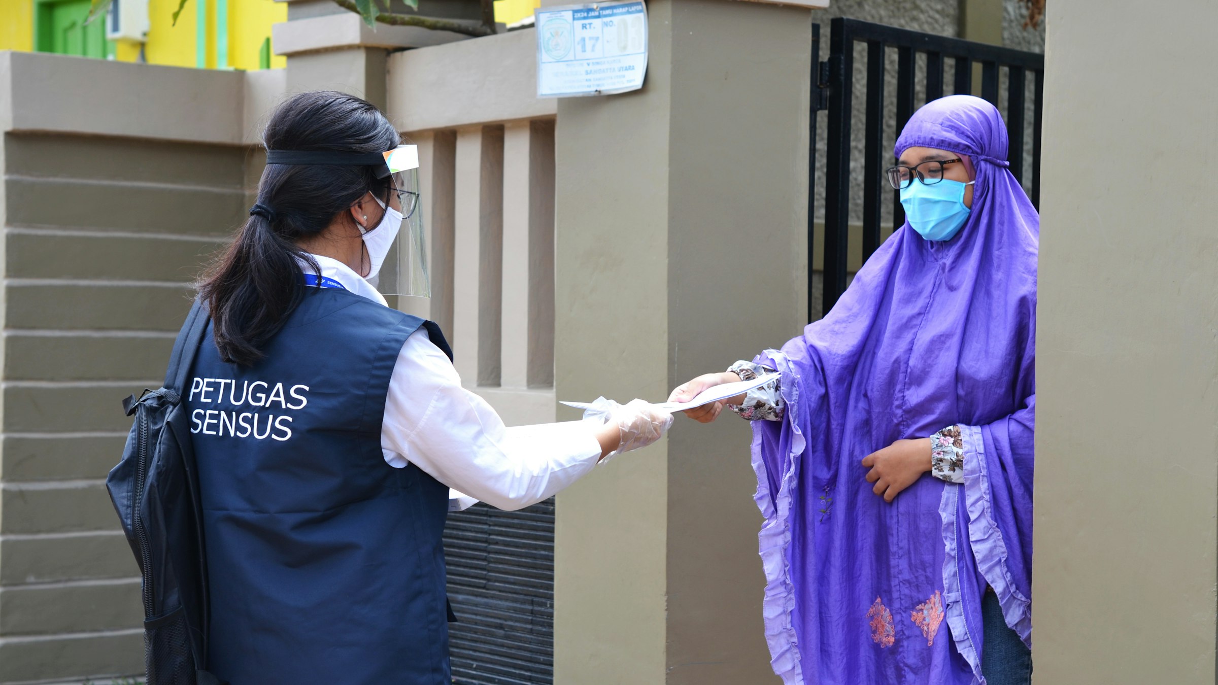 Census officer in PPE handing paperwork to a household member at her gate, Indonesia